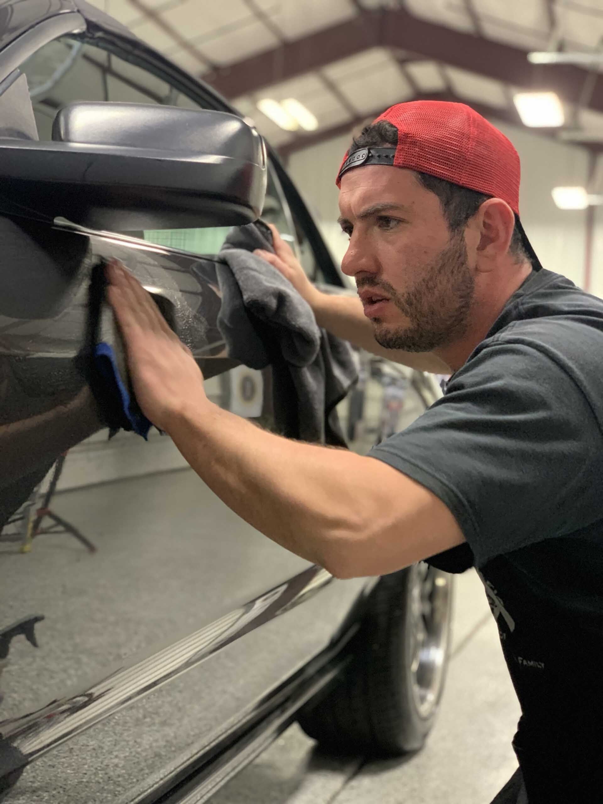 Man in red cap polishing a dark car door with a blue cloth inside a garage.