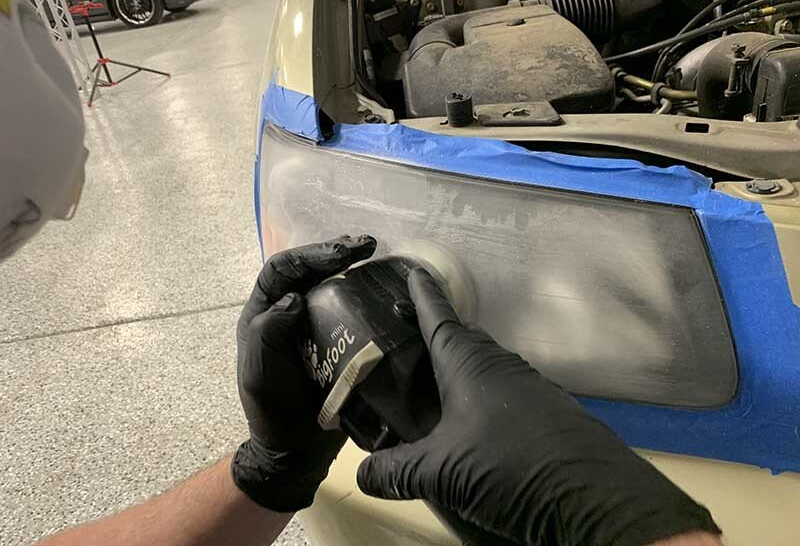 Person wearing black gloves sanding a car headlight with a power sander, exterior, blue tape.