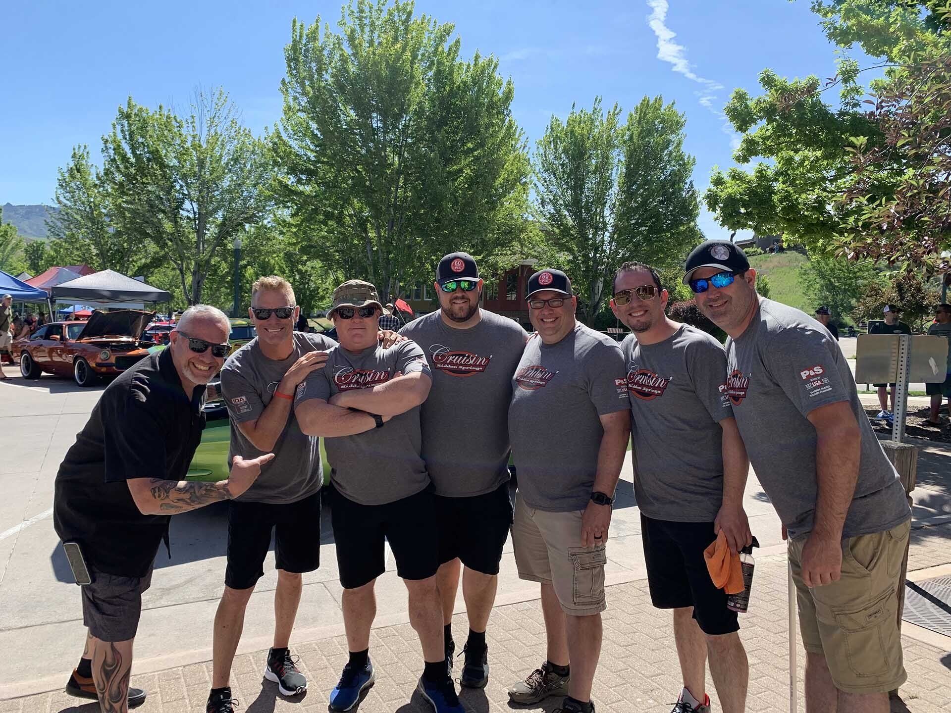 Group of seven men smiling, posing outside on a sunny day. They wear matching gray shirts with a logo.
