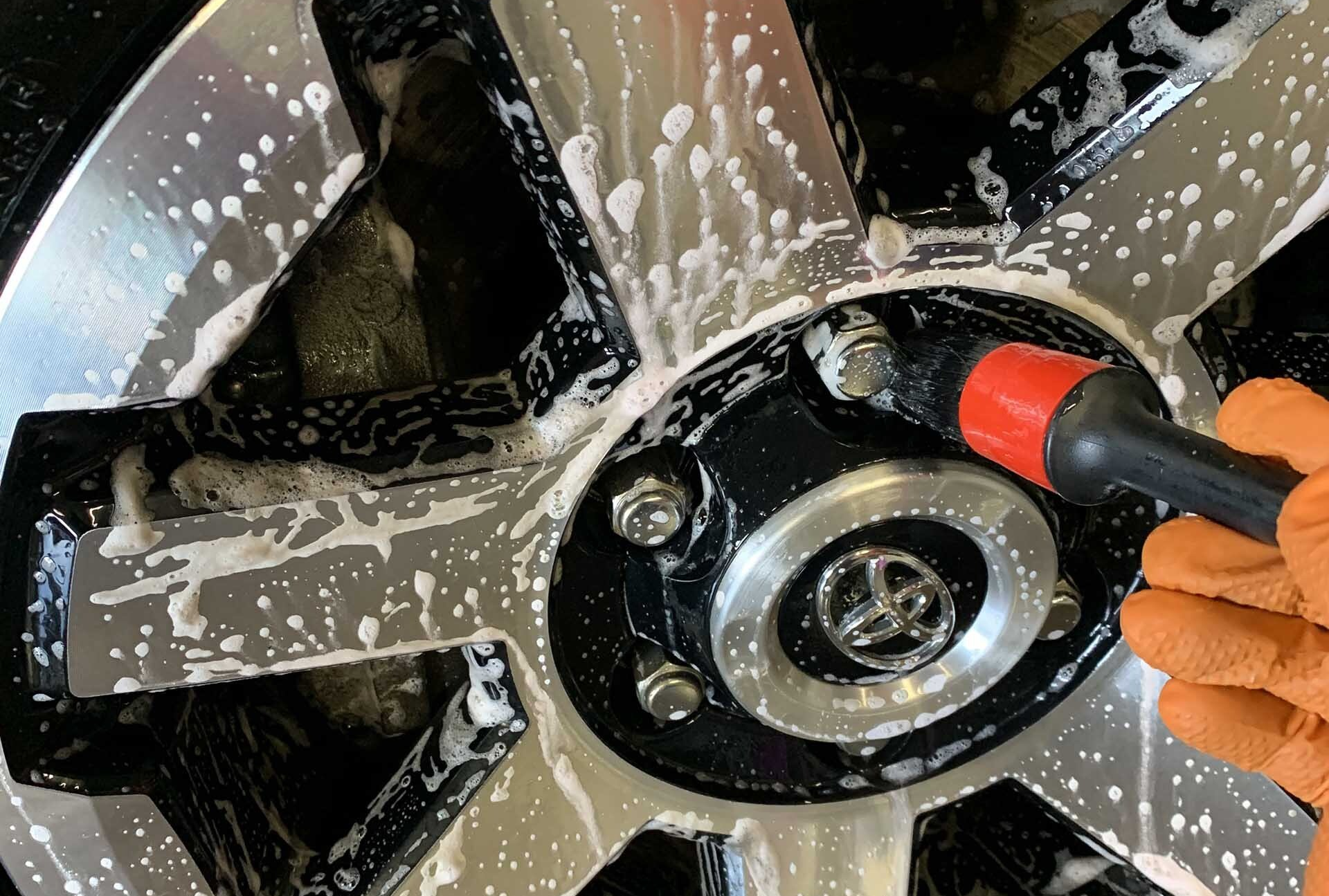 Person wearing orange gloves scrubs a soapy black and silver car wheel with a brush.