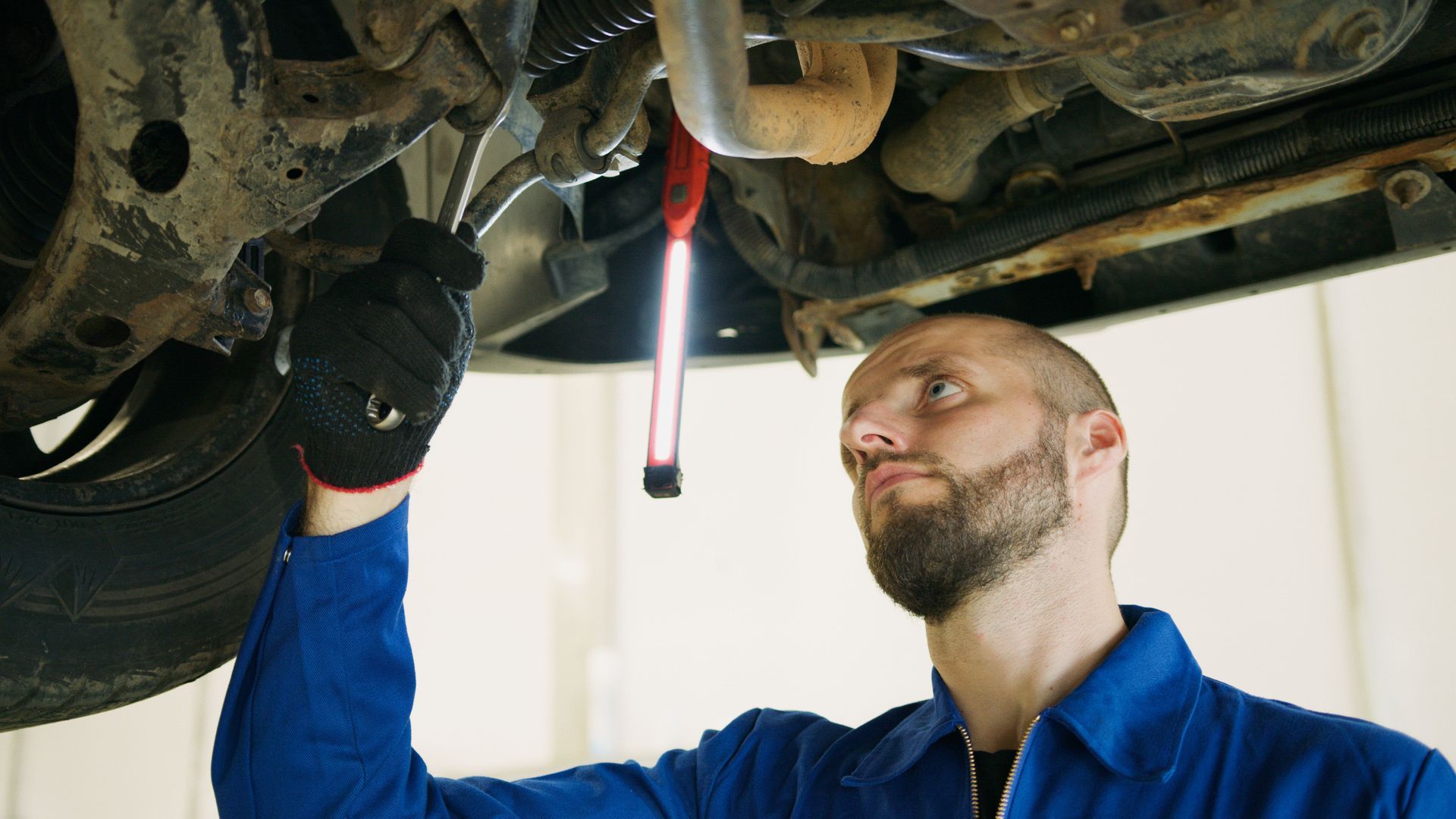 Mechanic in blue coveralls inspecting undercarriage of a car with a flashlight | HD Auto Care and More