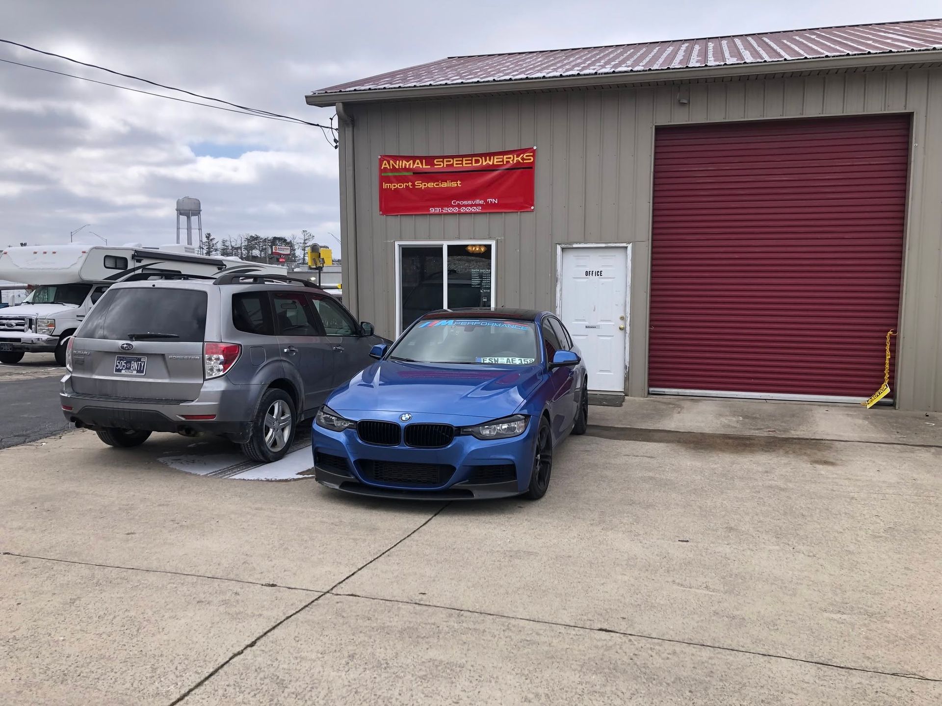 Blue sedan and gray SUV parked outside a garage with a red roll-up door and red sign.
