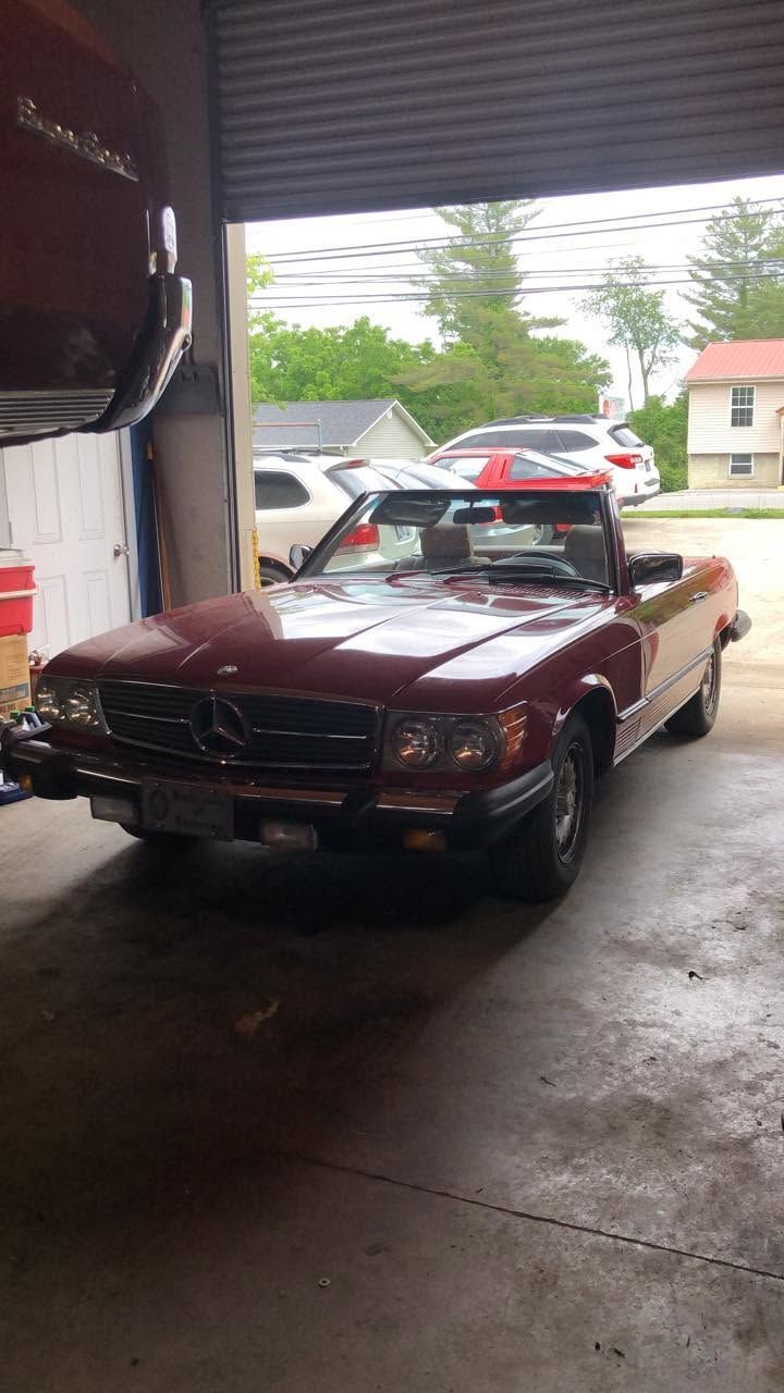Maroon vintage convertible parked inside a garage, with other cars visible outside.