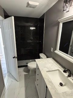 Modern bathroom with white vanity, marble countertop, black tiled shower, and a toilet beside a window.