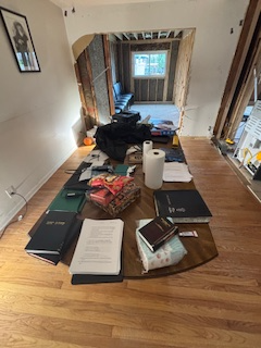 Hallway cluttered with books, bags, and household items on wooden floors, leading to a room with a window.