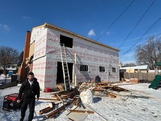House under construction with exposed framing, ladder, snow on the ground, and a worker in front