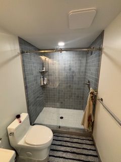 Narrow accessible bathroom with toilet, walk-in shower, gray tile, and striped bath mat