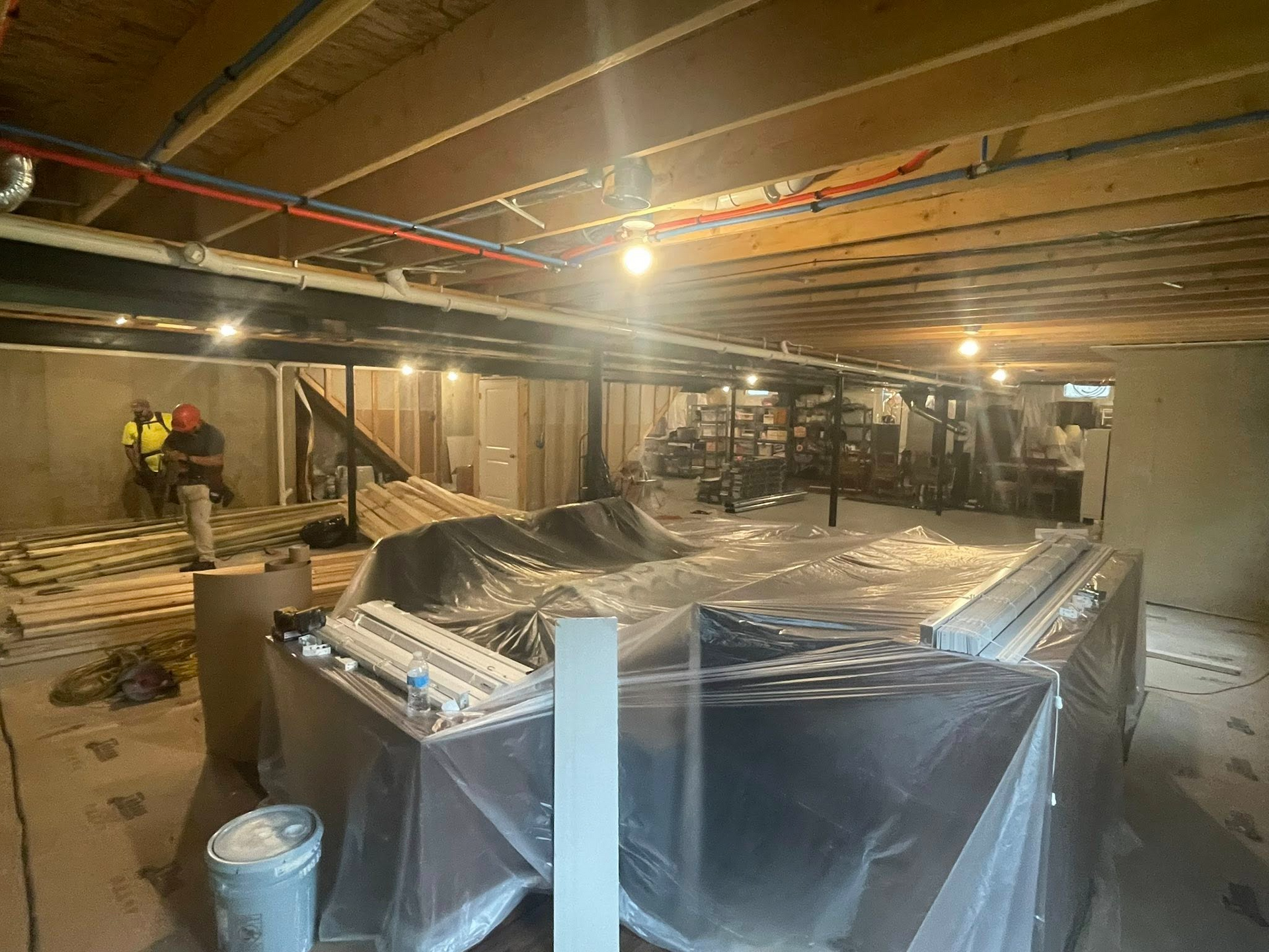 Basement under construction with workers, exposed ceiling pipes, and plastic-covered equipment.