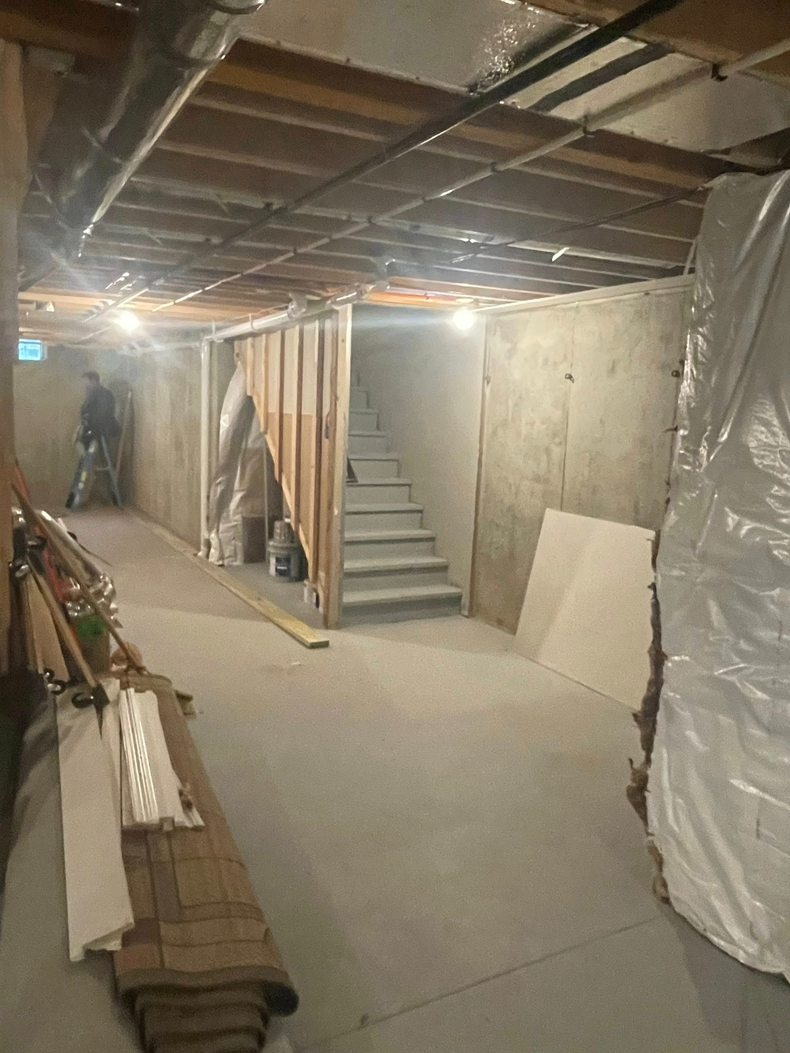 Unfinished basement with concrete floor, exposed ceiling, staircase, and stacked lumber along the wall.