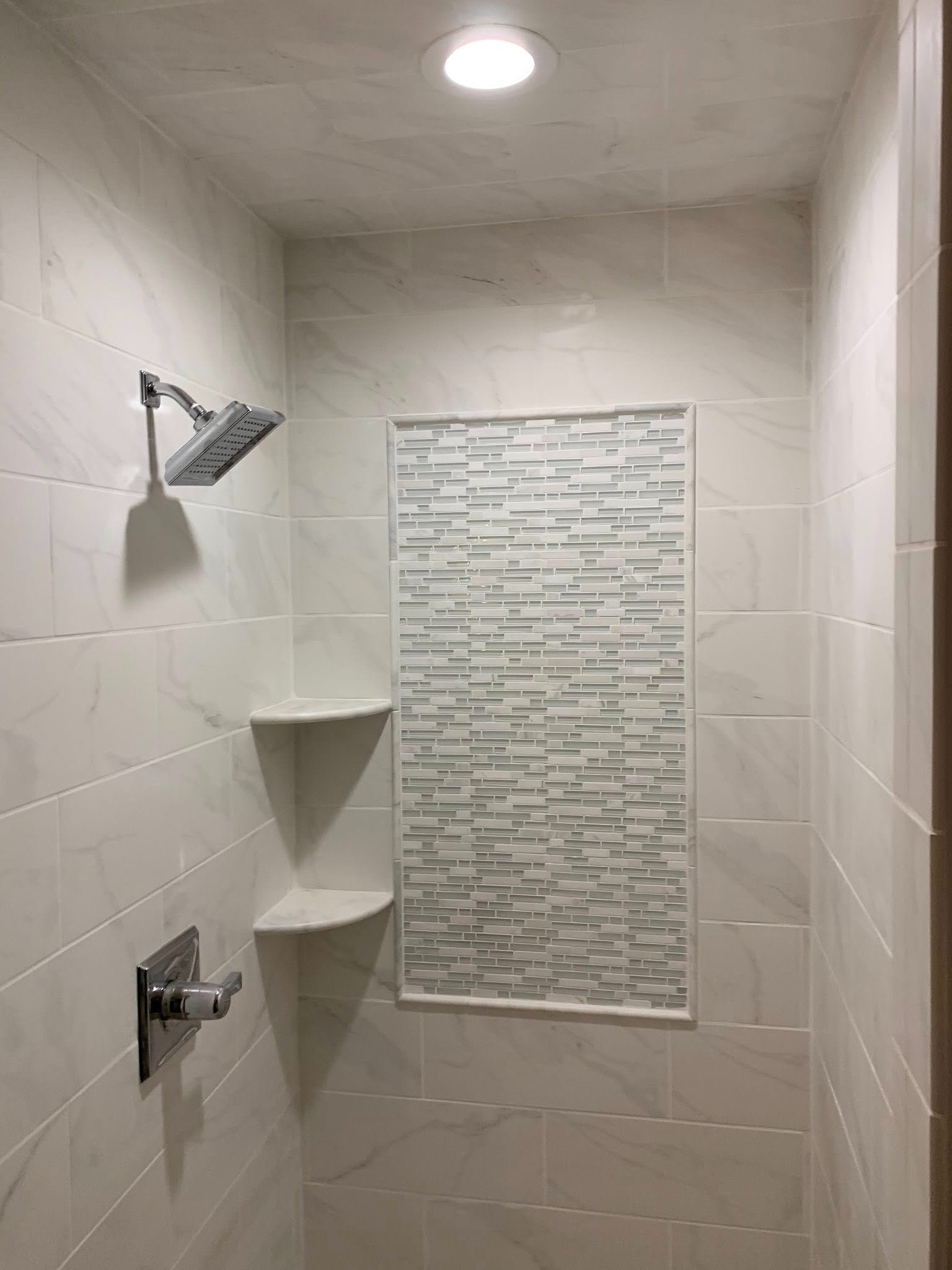 White tiled shower with two showerheads, corner shelves, and a decorative mosaic tile panel.