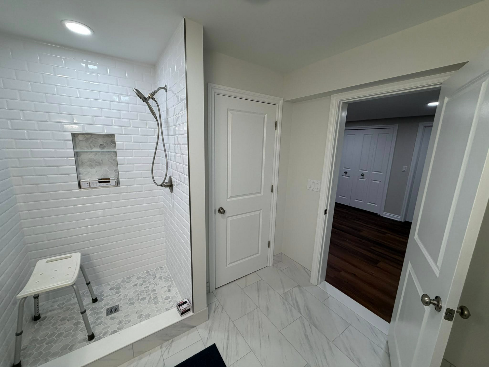 Bright bathroom with white tile shower and open door to a dark wood-floored room