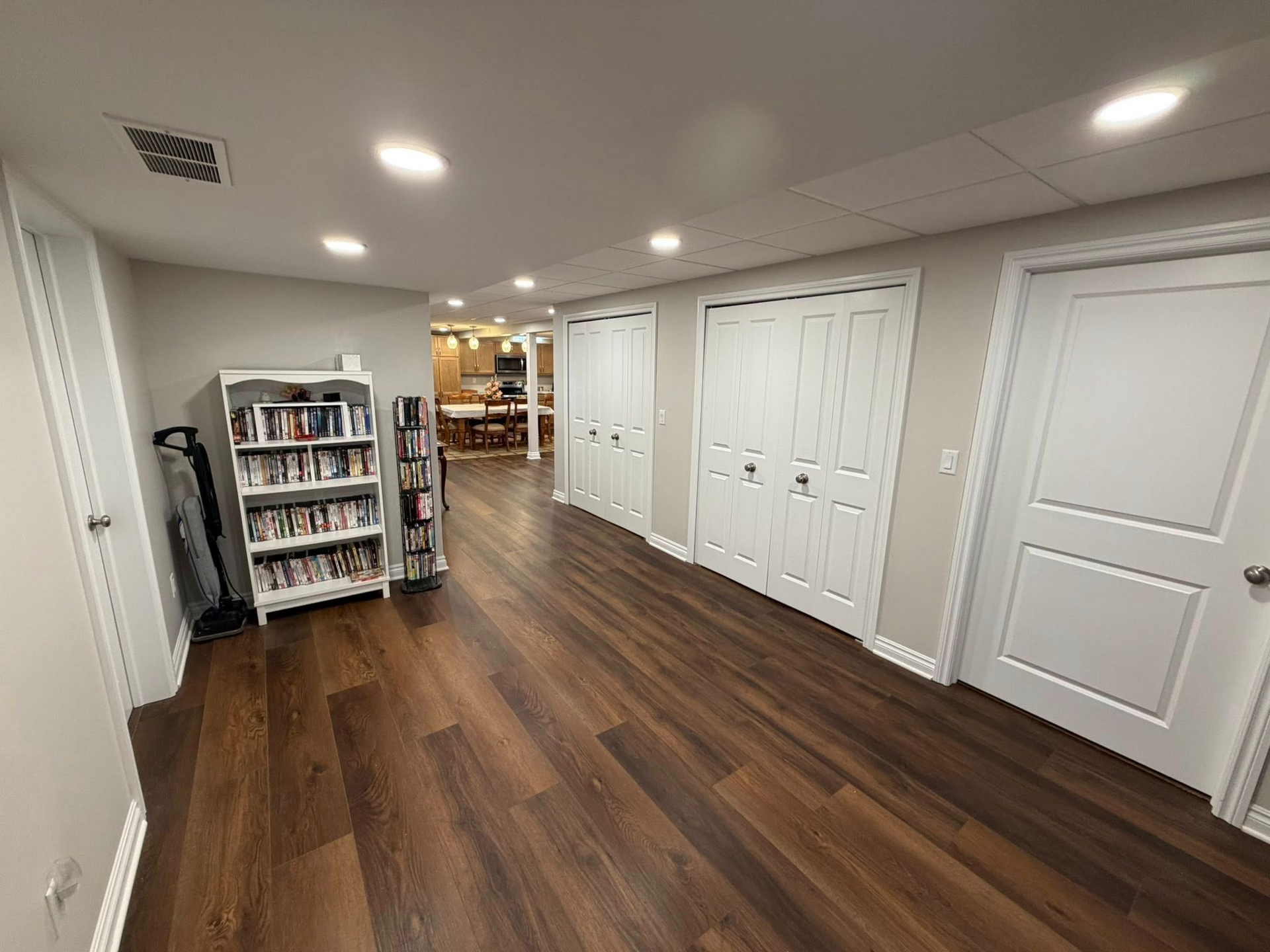 Empty hallway with wood floors, white doors, recessed lights, and a bookshelf near the back.