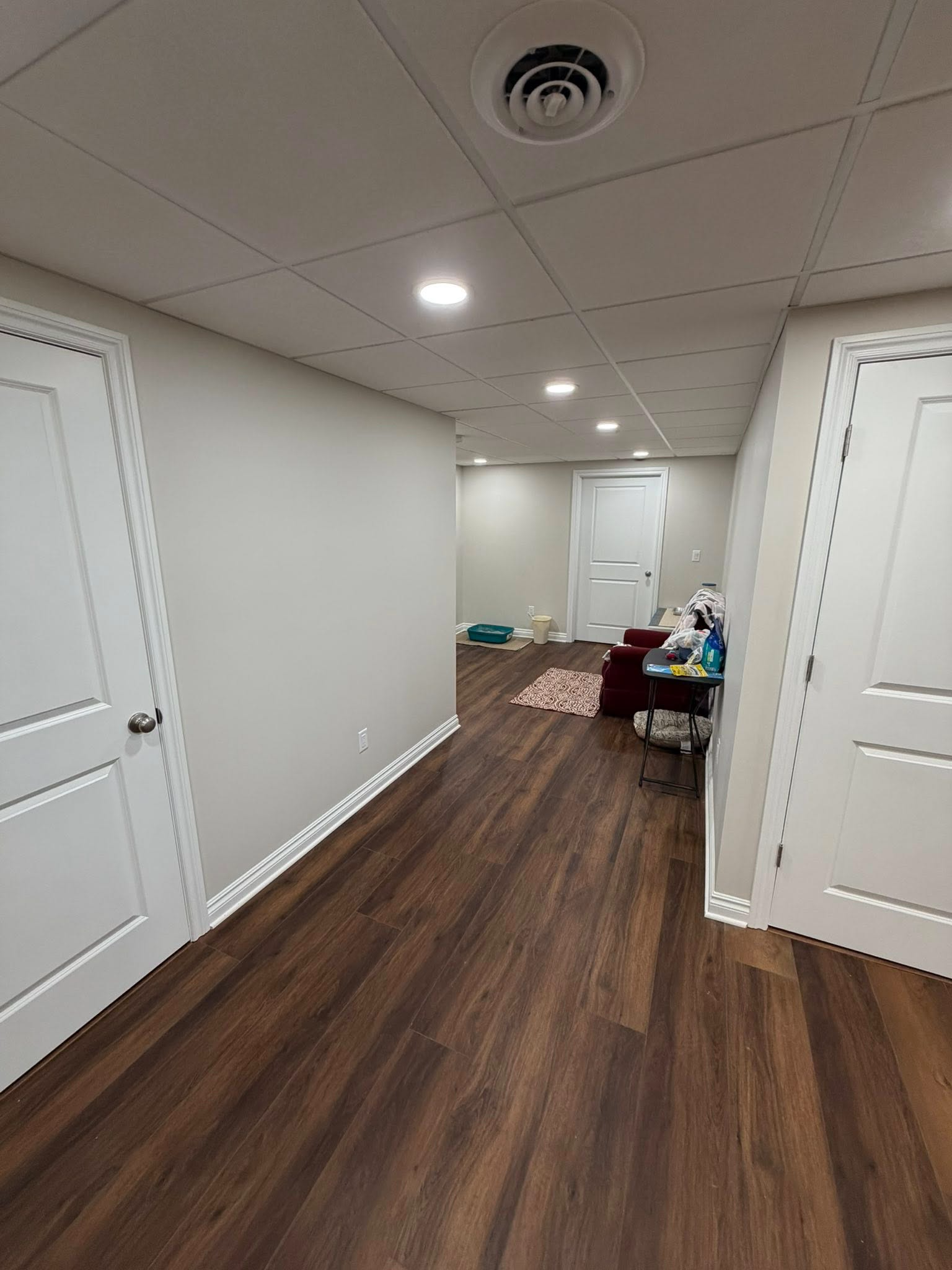 Empty hallway with white walls and doors, wood flooring, and a lit room at the end.