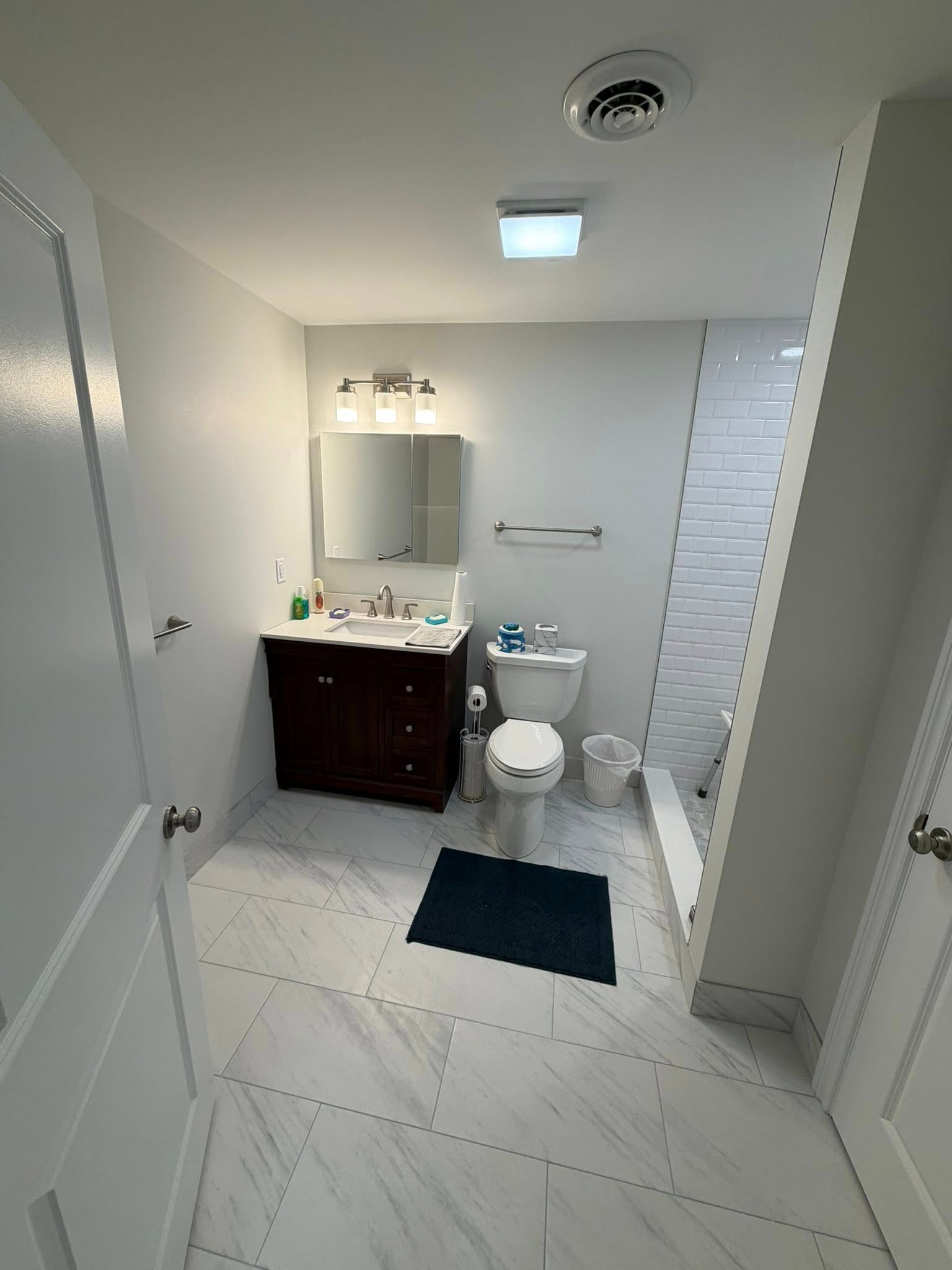 Small bathroom with white tile floor, dark vanity, toilet, mirror, and shower stall on the right.