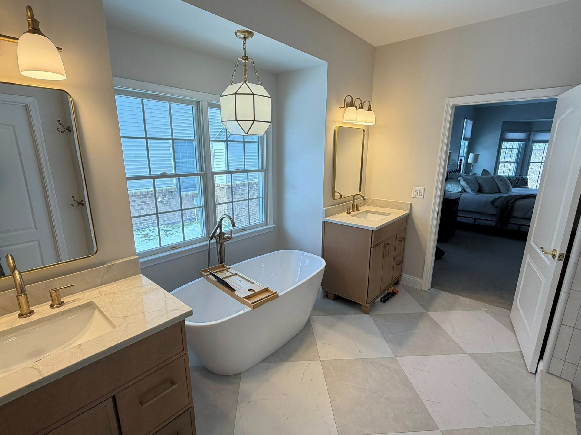 Bright bathroom with freestanding tub by window, double vanity, and light tile floor leading to bedroom view