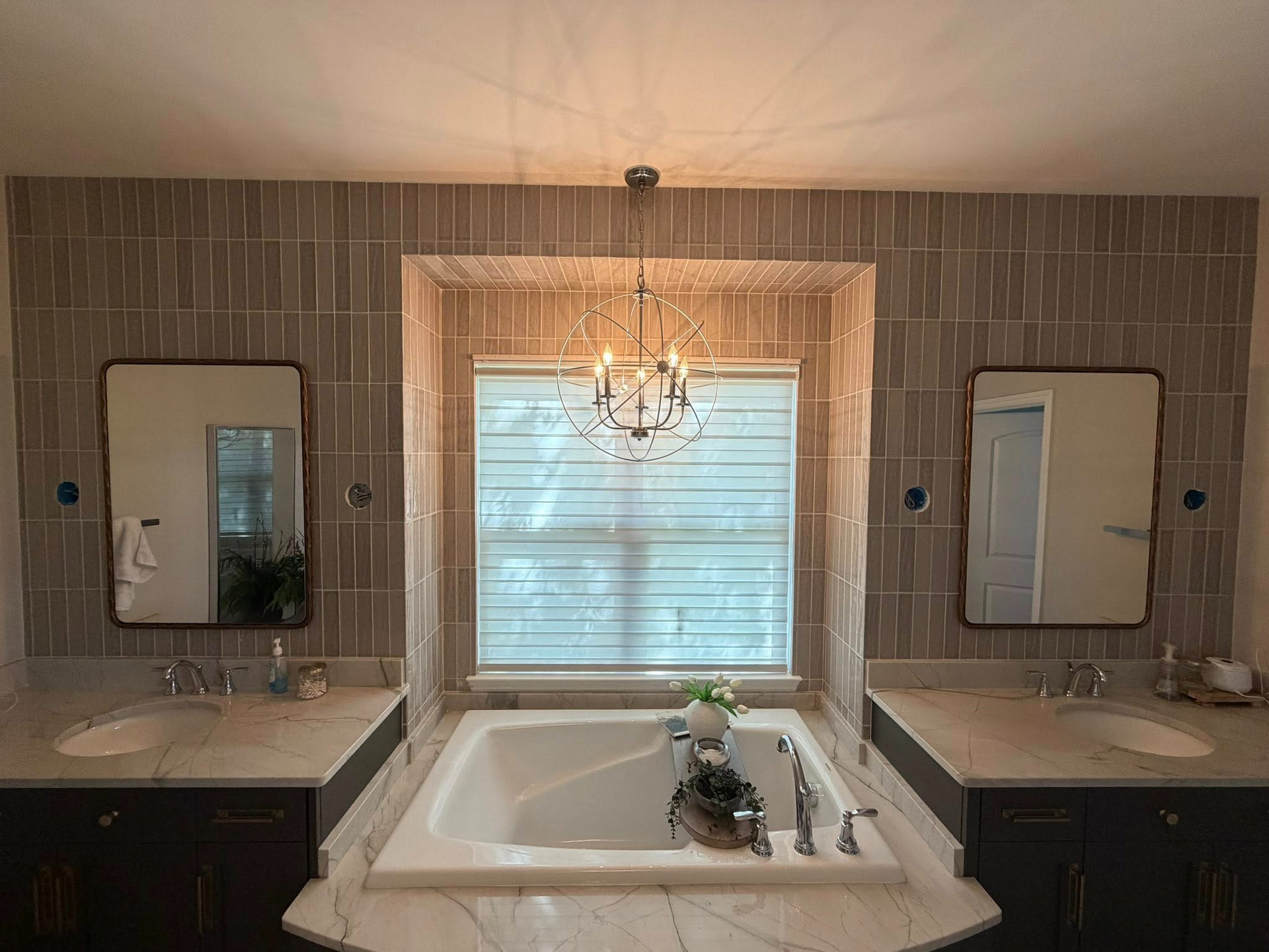 Luxury bathroom with double sinks, large central bathtub, tiled walls, and a chandelier above the window.