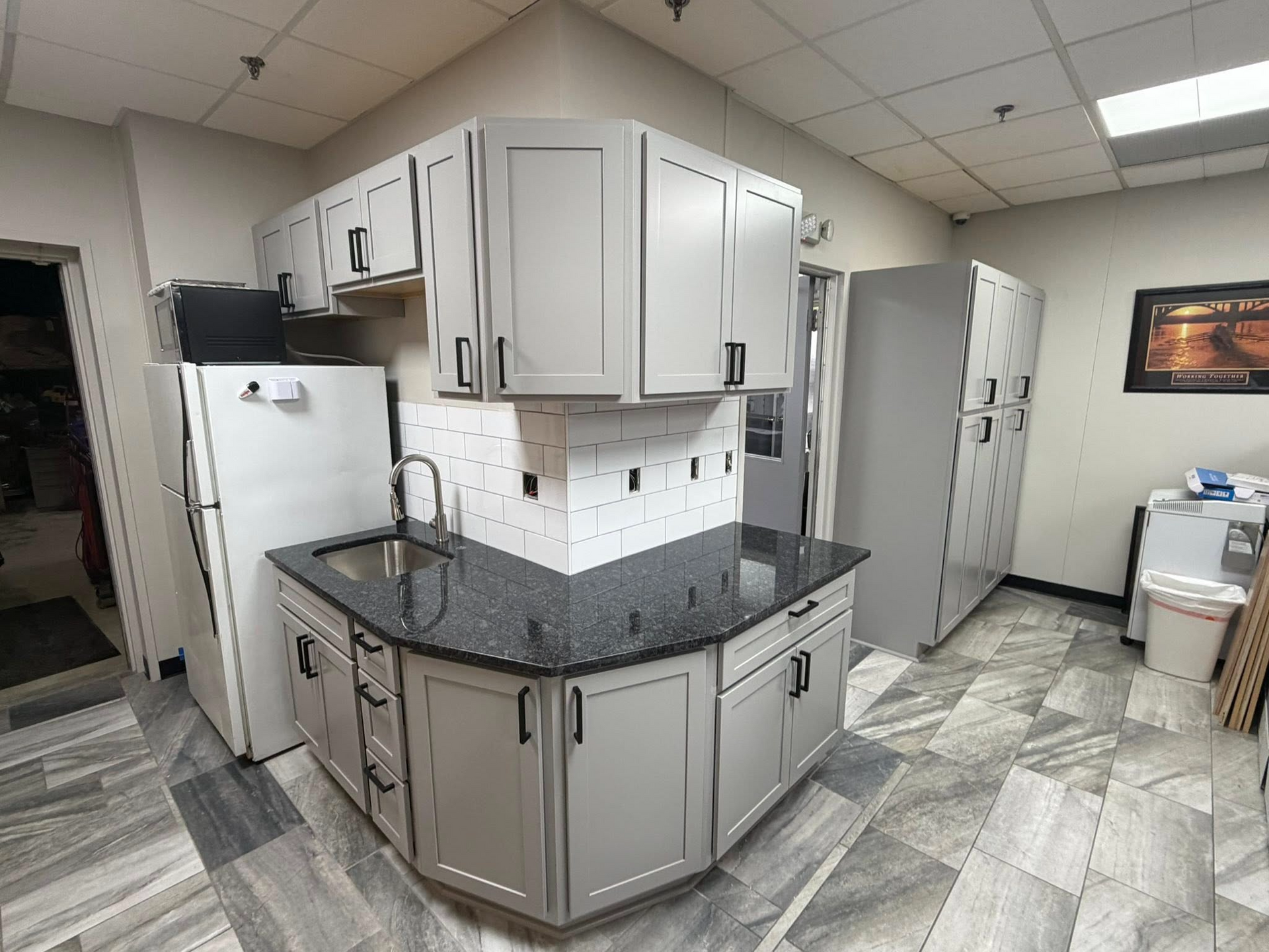 Office break room with gray cabinets, black countertops, refrigerator, and tiled floor