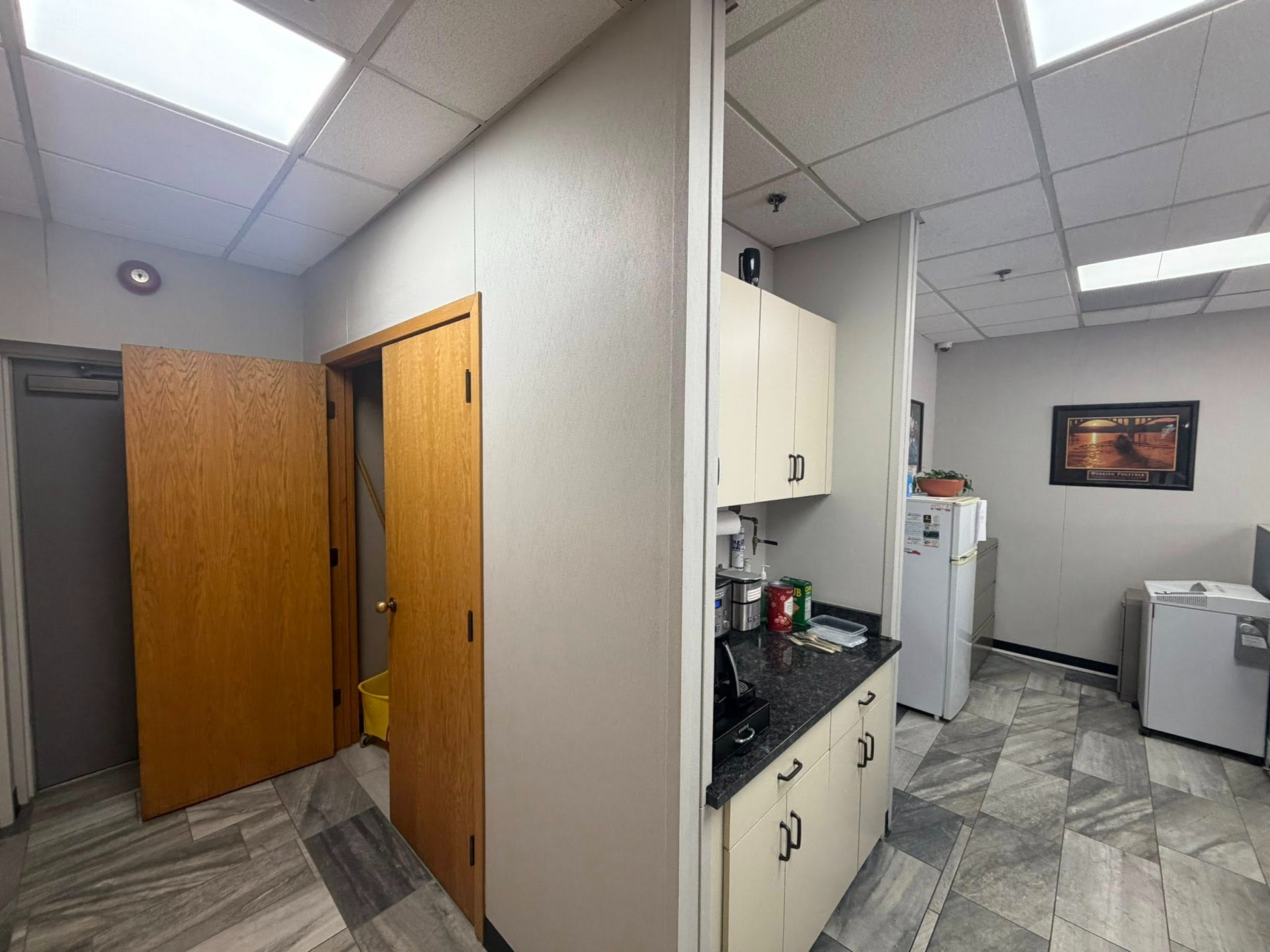 Office break room with kitchenette, white cabinets, refrigerator, sink, and open wooden door.
