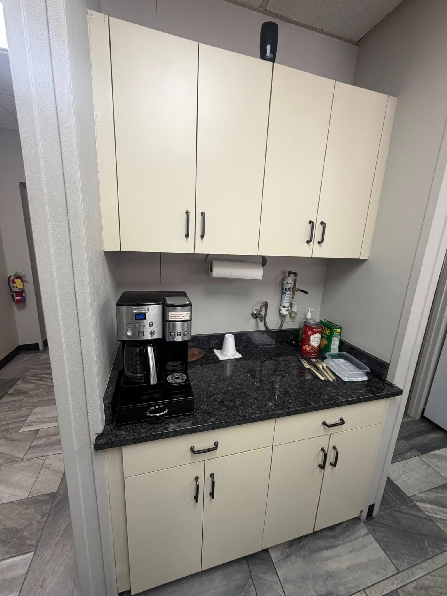 Small kitchenette with cream cabinets, black countertop, coffee maker, sink, and cup dispenser.