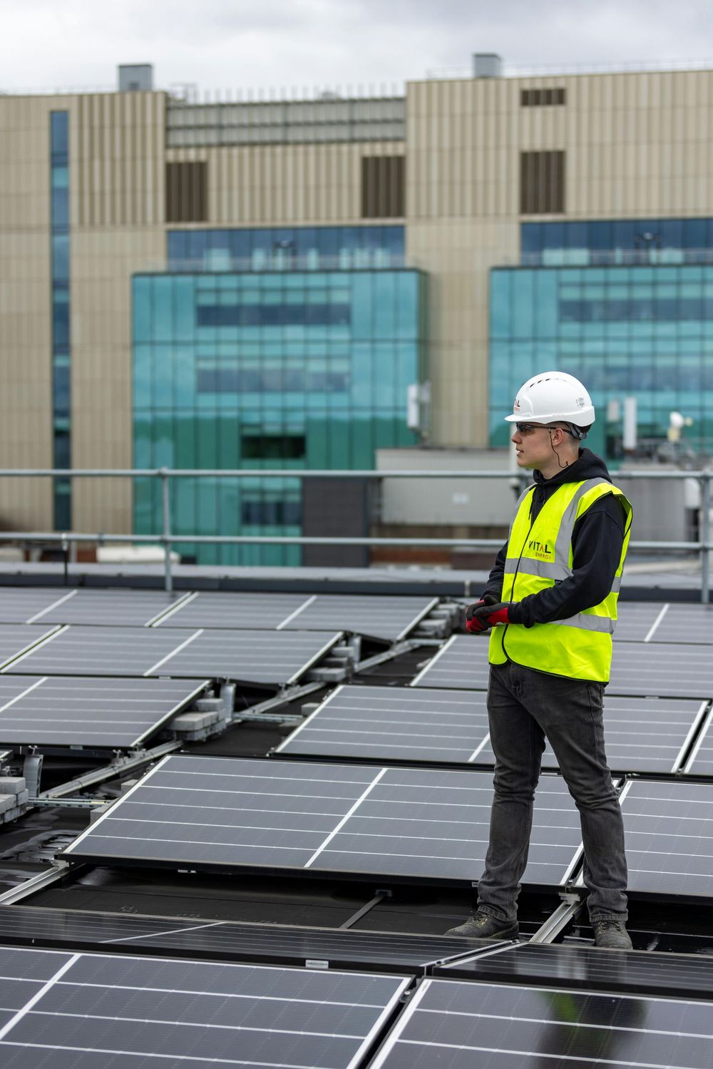 Worker in hard hat standing on rooftop solar panels with a modern building in the background