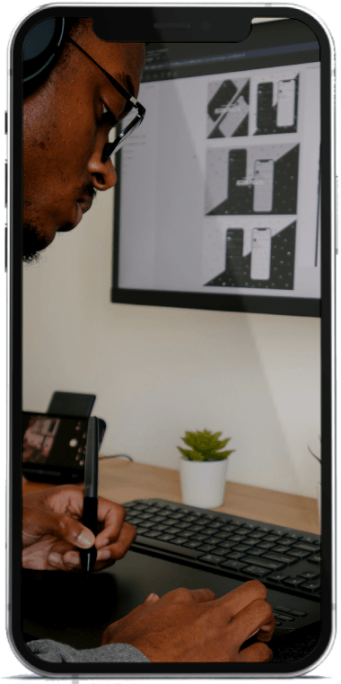 A person using a stylus at a desk with a screen showing designs, a phone, headphones, and a plant.