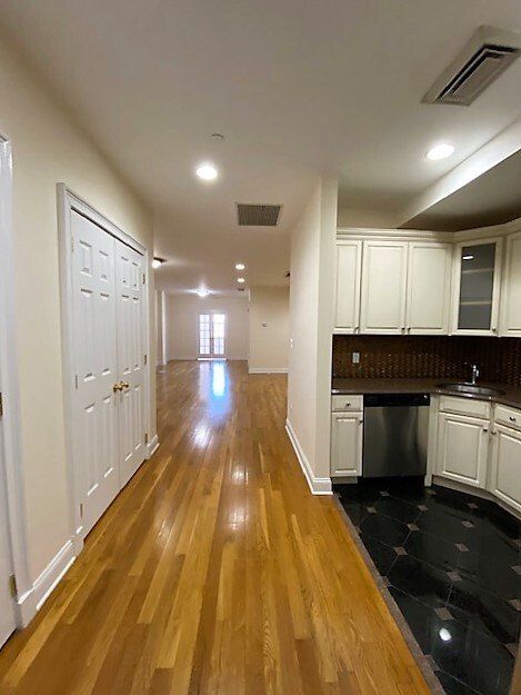 A hallway with light wood floors leads past a kitchen with white cabinets, dark counters, and black tiled flooring.