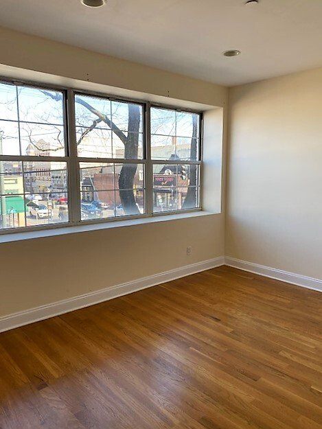 A bright, empty room with light walls, hardwood floors, and a large window overlooking a city street.