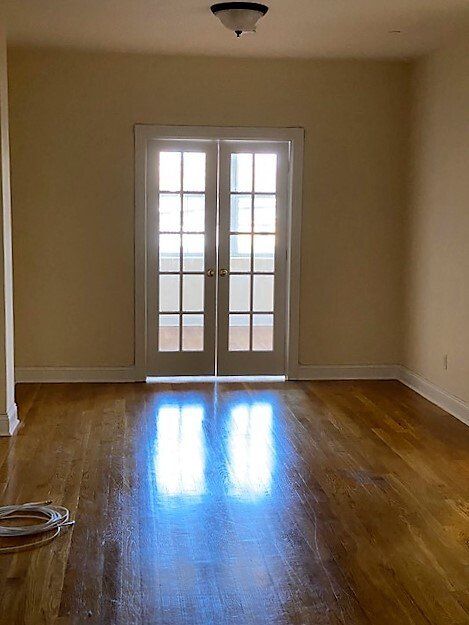 An empty room with polished hardwood floors, cream-colored walls, and a pair of French doors leading outside.
