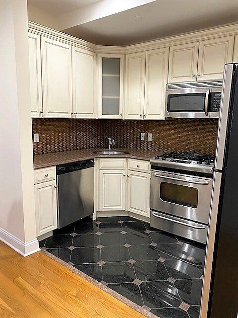 Kitchen with cream-colored cabinets, stainless steel appliances, dark tile floor, and brown backsplash.