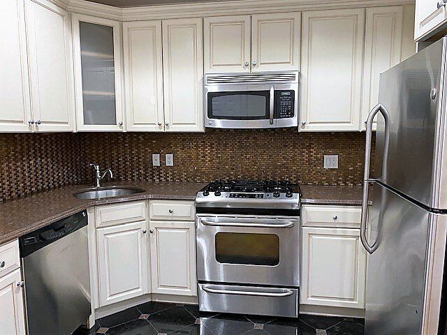 A modern kitchen with off-white cabinetry, stainless steel appliances, dark countertops, and a textured brown backsplash.