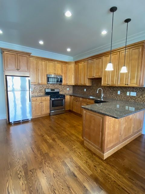 A kitchen with light wood cabinets, stainless steel appliances, granite countertops, an island, and dark wood flooring.