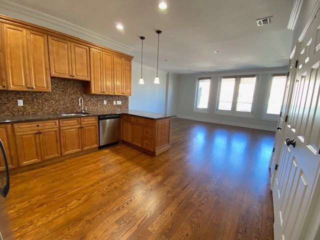 A kitchen with light wood cabinets, a brown tiled backsplash, and hardwood floors leading into an open living space.
