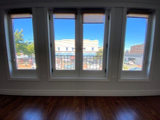 Four tall, white-framed windows look out onto a sunny street scene with buildings and trees, above a wood floor.