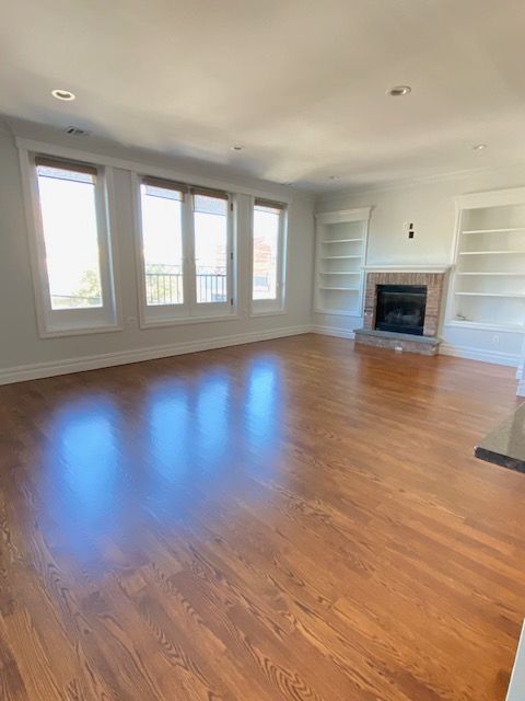A spacious, light-filled room with polished hardwood floors, large windows, and a brick fireplace flanked by bookshelves.