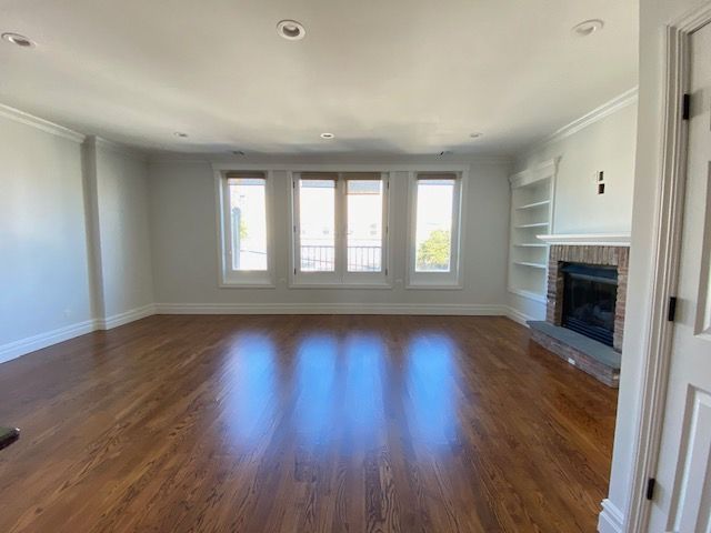 A bright, empty living room with hardwood floors, a stone fireplace, built-in shelving, and large windows.