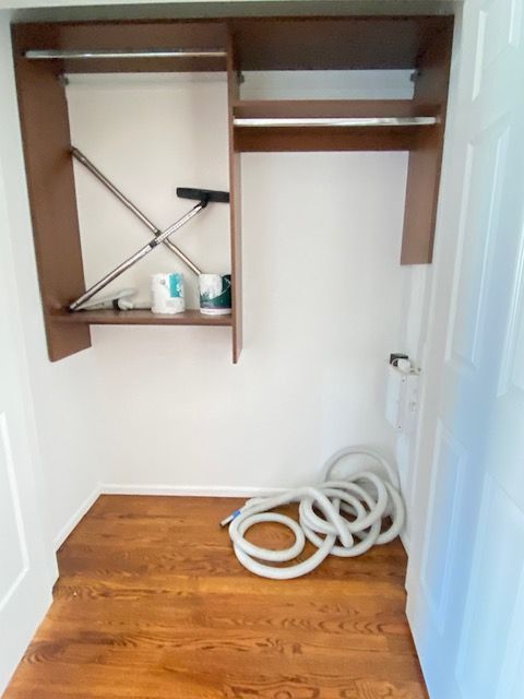 An open walk-in closet with brown wooden shelves, a hanging rod, a cleaning tool, supplies, and a coiled vacuum hose.