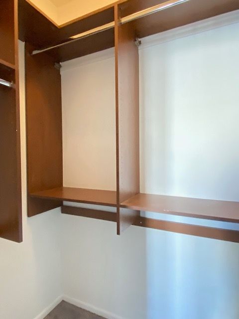 A walk-in closet interior with brown wooden shelves, white walls, and empty metal clothes hanging rods.