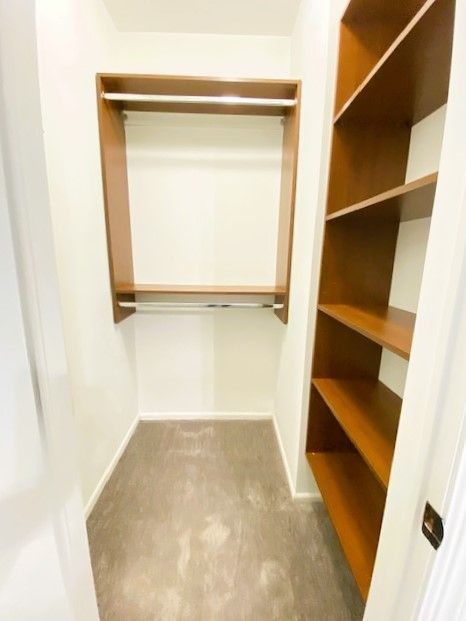 A small, empty walk-in closet with light grey flooring, a wooden hanging rod, and built-in wooden shelving on one side.