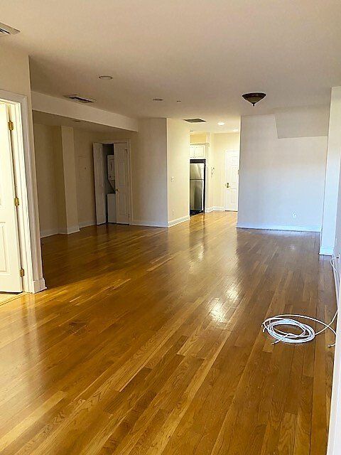 A wide, empty room with polished hardwood floors, recessed lighting, and a view into a kitchen with stainless appliances.