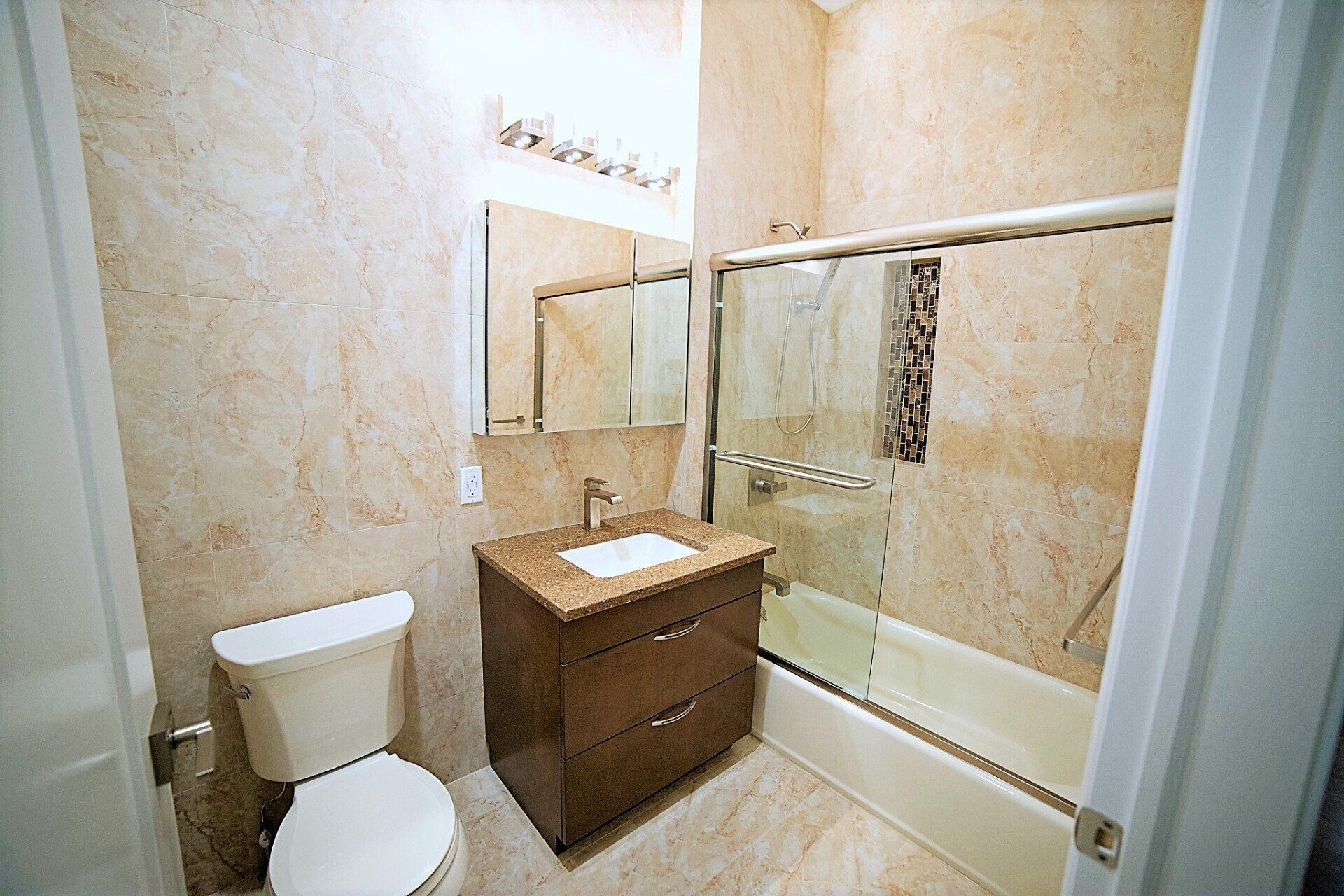 A bathroom with tan marble-style walls, a brown vanity, a toilet, and a glass-enclosed shower/tub combination.