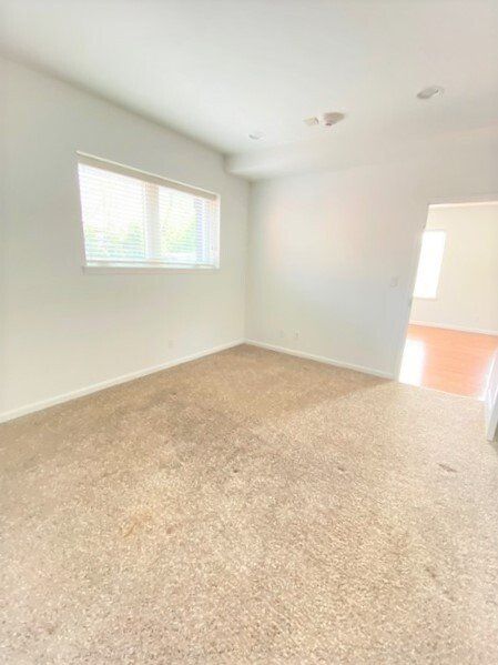 An empty room with beige carpet, white walls, a single window on the left, and an open doorway leading to another room.