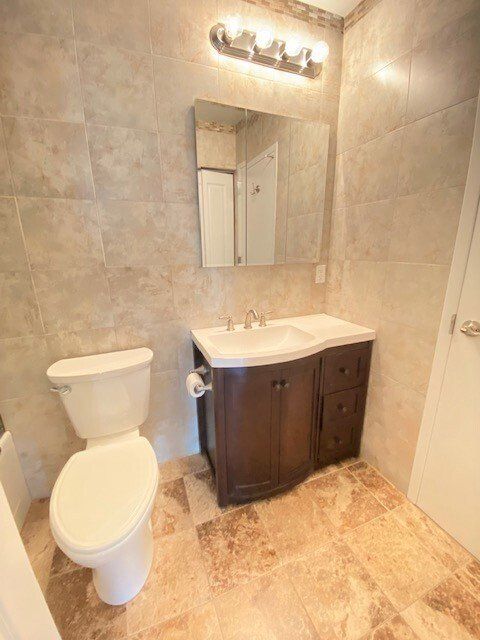 A bathroom with a white toilet, dark wood vanity, stone tile walls and floors, and a modern rectangular vanity light.