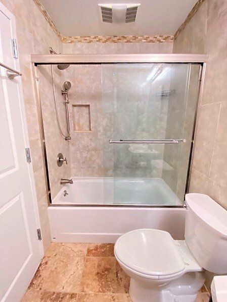 A bathroom with a white toilet in the foreground, a tub-shower combo with glass doors, and tan tiled walls and flooring.