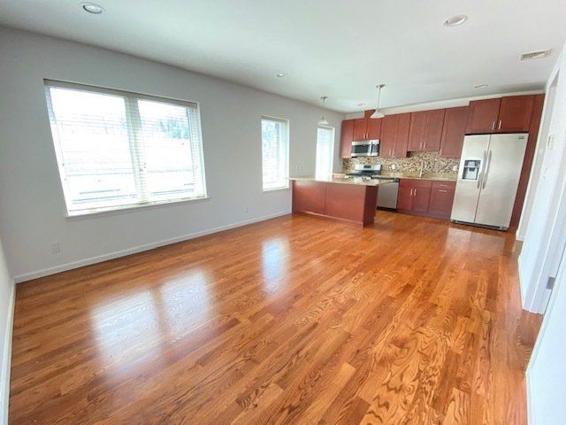 An open-plan room with polished hardwood floors, light gray walls, recessed lighting, and a view into a wooden kitchen.