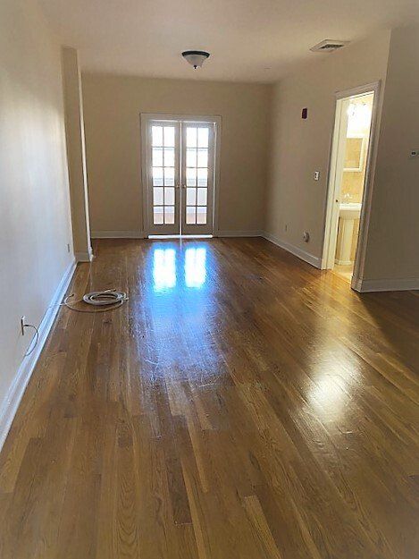 An empty room with glossy hardwood floors, beige walls, a French door, and an open doorway leading to a small bathroom.
