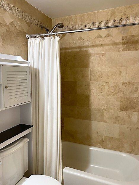 A bathroom shower with a white curtain, a chrome shower head, and beige stone-tiled walls above a white bathtub.