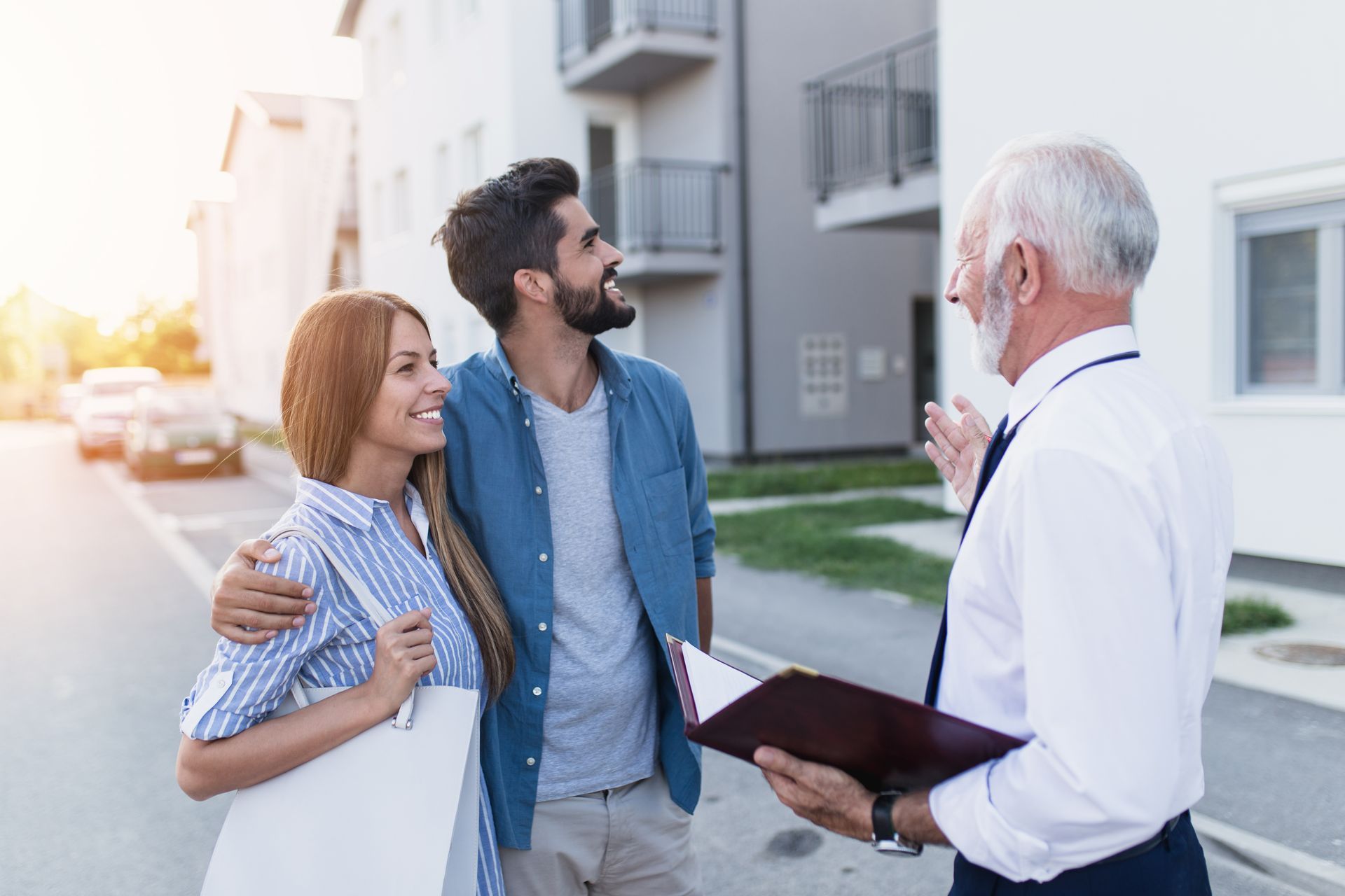 A senior agent presents a real estate rental property to a smiling young couple at sunset.