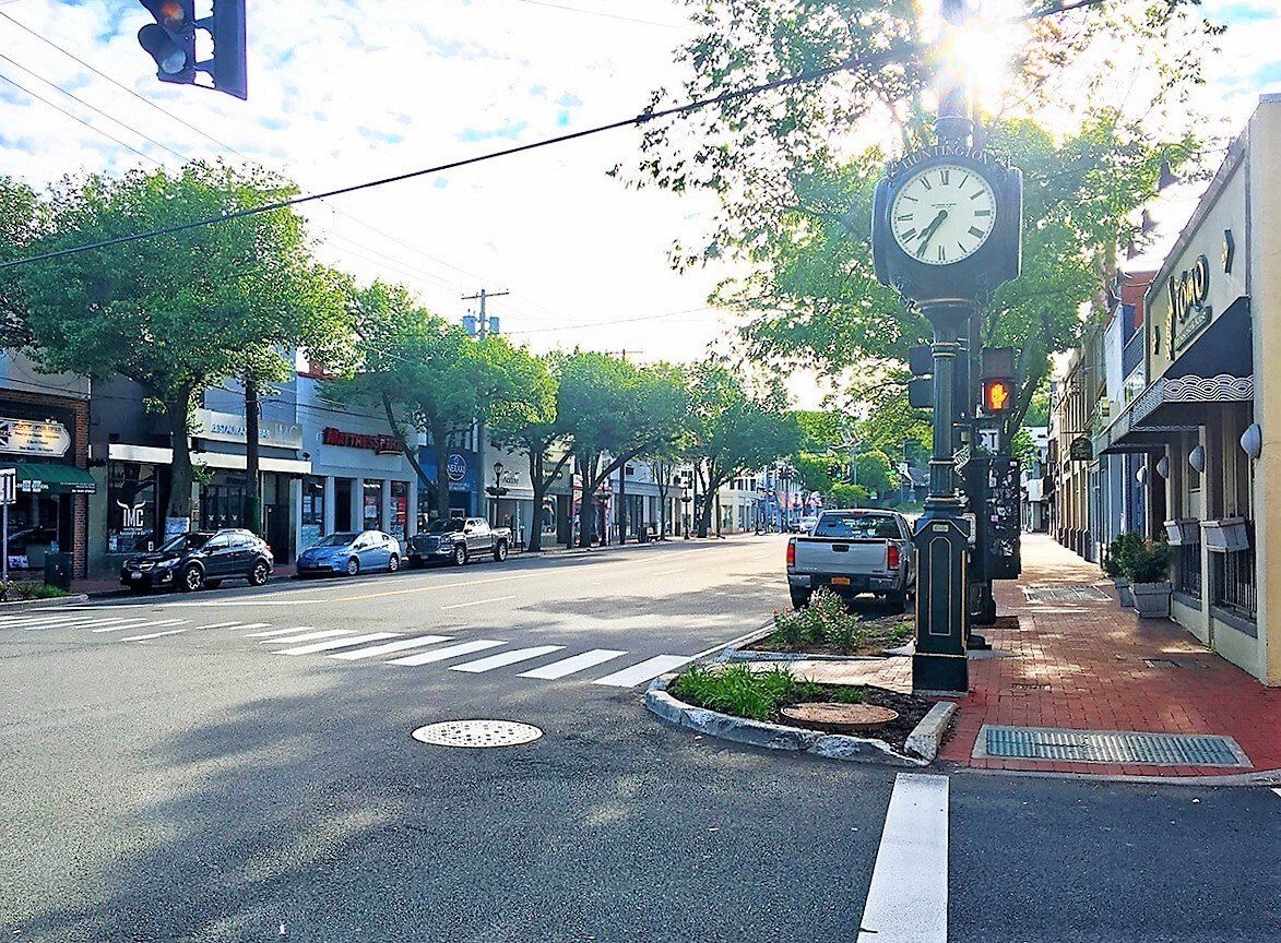 A street-level view of a sunlit downtown street lined with trees, storefronts, and a prominent sidewalk clock.