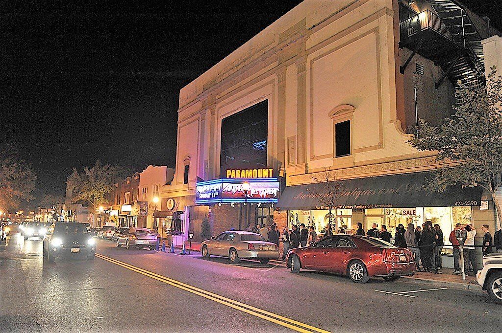A night scene of a historic theater on a street with parked cars and a group of people gathered outside.