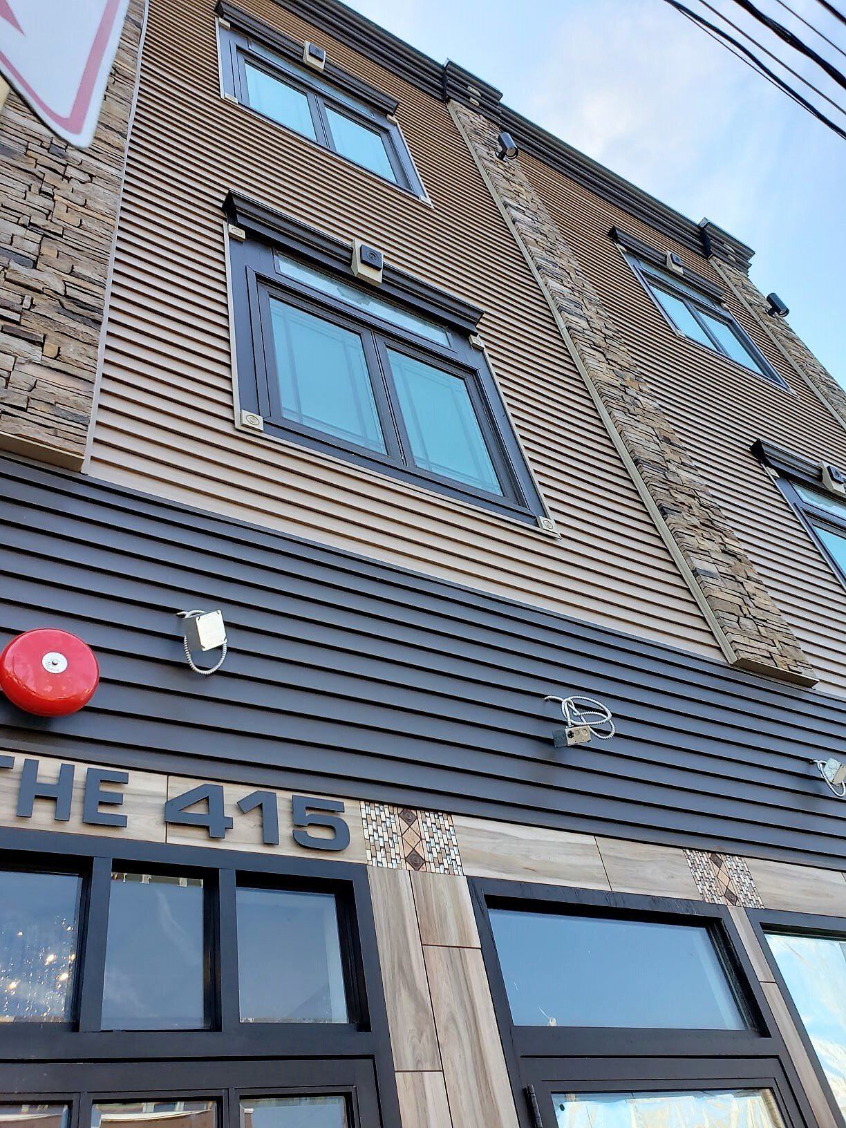 A low-angle view of a building facade with grey horizontal siding, windows, decorative trim, and a red fire alarm bell.